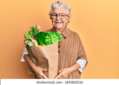 Senior grey-haired woman holding paper bag with bread and groceries looking positive and happy standing and smiling with a confident smile showing teeth  - Powered by Shutterstock