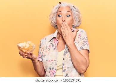 Senior Grey-haired Woman Holding Potato Chip Covering Mouth With Hand, Shocked And Afraid For Mistake. Surprised Expression 