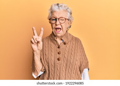Senior grey-haired woman wearing casual clothes and glasses smiling with happy face winking at the camera doing victory sign. number two. 