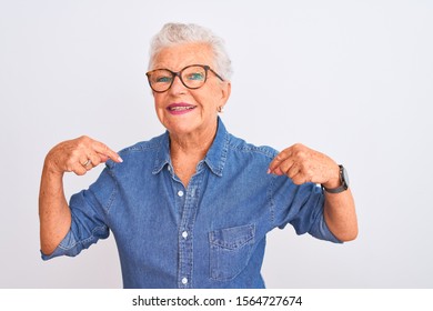Senior grey-haired woman wearing denim shirt and glasses over isolated white background looking confident with smile on face, pointing oneself with fingers proud and happy.