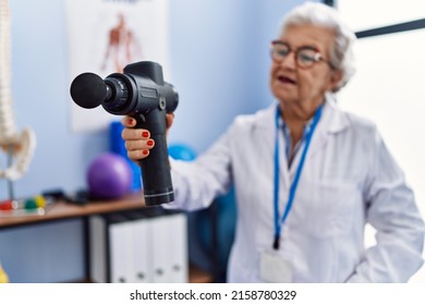 Senior Grey-haired Woman Wearing Physiotherapist Uniform Holding Percussion Gun At Physitherapy Clinic