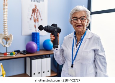 Senior Grey-haired Woman Wearing Physiotherapist Uniform Holding Percussion Gun At Physitherapy Clinic