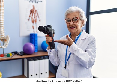 Senior Grey-haired Woman Wearing Physiotherapist Uniform Holding Percussion Gun At Physitherapy Clinic