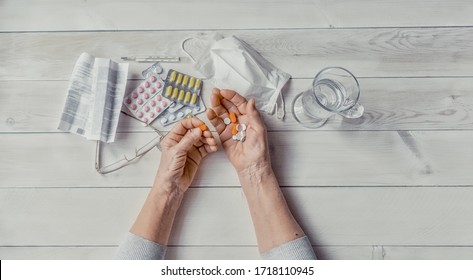 Senior Hands, Pills And Drugs On Table, Glass Of Water. Wrinkled Hands Of Old Woman Holding Colorful Tablets, Mask, Glasses, Prescription, Thermometer, Wooden Background.  Health Care For Elderly.