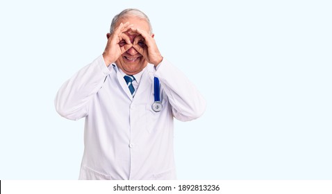 Senior handsome grey-haired man wearing doctor coat and stethoscope doing ok gesture like binoculars sticking tongue out, eyes looking through fingers. crazy expression. 
