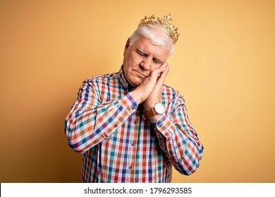 Senior handsome hoary man wearing golden crown of king over isolated yellow background sleeping tired dreaming and posing with hands together while smiling with closed eyes.