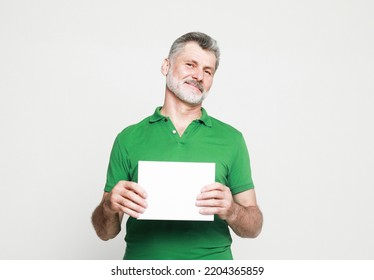 Senior Handsome Man With Mustache And Beard Wearing Green Shirt Holds The White Sign In A Studio Over Light Grey Background. Look Here, Great Offer.