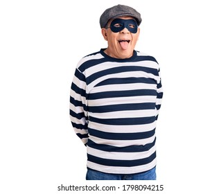 Senior Handsome Man Wearing Burglar Mask And T-shirt Sticking Tongue Out Happy With Funny Expression. Emotion Concept. 