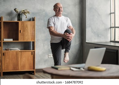 Senior handsome sportman doing stretching and balancing yoga exercise at home using online lesson on notebook. How to stay healthy on quarantine concept. - Shutterstock ID 1684787092