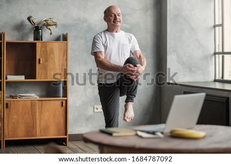 Senior handsome sportman doing stretching and balancing yoga exercise at home using online lesson on notebook. How to stay healthy on quarantine concept. Сток-фото © 