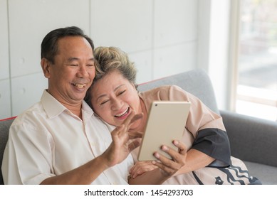 Senior Happy Asian Couple Sitting On Couch And Using Smart Tablet For Facetime, Online Shopping, Order Food Delivery Or Media Entertainment In Modern Living Room At House. Social Distancing.