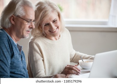 Senior Happy Couple Enjoying Using Laptop Eating Breakfast Together, Smiling Elderly Middle Aged Family Shopping Online, Reading Discussing Internet News, Buying Travel Tour On Computer Site At Home