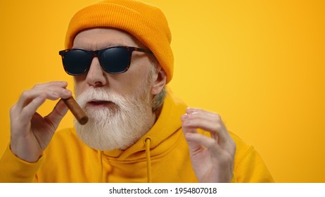 Senior happy man putting cigar to mouth on modern background. Satisfied old guy enjoying smell of cigar indoors. Successful aged male person relaxing on yellow studio background.