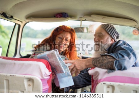 A senior hipster couple sitting in a car, drinking coffee and looking at a road map. he is tattooed and he wears a white beard,
