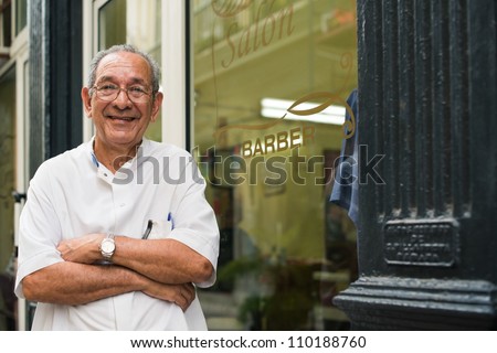 senior hispanic barber in old fashion barber's shop, posing and looking at camera with arms crossed near shop window