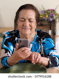 Senior Hispanic Or Latina Woman Holding And Using Her Smartphone While Sitting Down