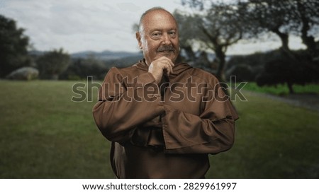 Senior hispanic man monk in brown habit touches chin with hand, folded arms and slight smile in forest field by trees and path; contemplation.