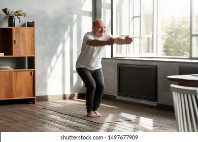 Senior Hispanic Man Practicing Yoga Indoors At Living Room Doing Chair Pose Or Utkatasana