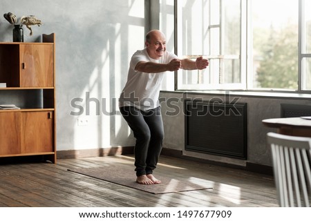 Senior hispanic man practicing yoga indoors at living room doing Chair pose or Utkatasana