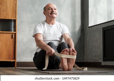 Senior Hispanic Man Sitting In Paschimottanasana Or Intense Dorsal Stretch Pose, Seated Forward Bend Posture, Exercise For Hips And Spine At Home Using Belt.