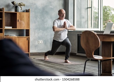 Senior Hispanic Man Standing In Warrior Yoga Pose Variation Practicing In Living Room Alone