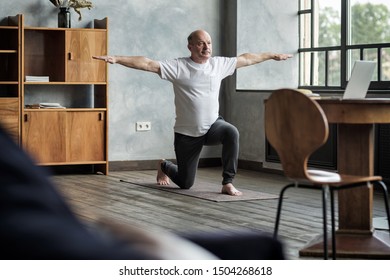 Senior Hispanic Man Standing In Warrior Yoga Pose Variation Practicing In Living Room Alone