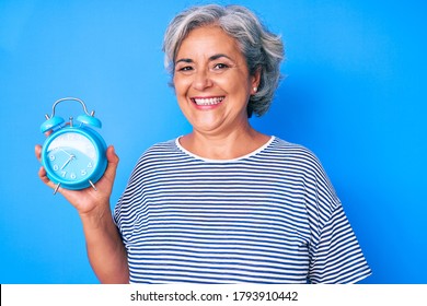 Senior hispanic woman holding alarm clock looking positive and happy standing and smiling with a confident smile showing teeth  - Powered by Shutterstock