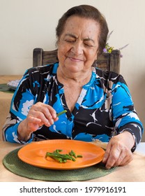 Senior Hispanic Woman Holding A Green Bean That She Picked From An Orange Plate