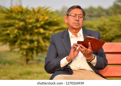 Senior Indian Man Curiously Checking And Working On His Smart Phone While Sitting On A Red Bench In A Park In Delhi, India