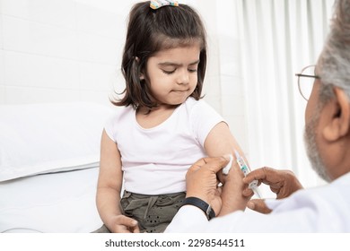 Senior Indian man pediatrician performs a vaccination of a little girl. Doctor administering an injection to a female child in a clinic. Injecting syringe needle in shoulder. - Powered by Shutterstock