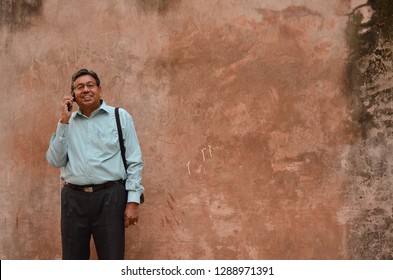 Senior Indian Man Smiling And Speaking On Phone Standing Against Brown Colored Wall Background In New Delhi, India