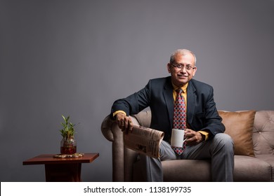 Senior Indian/asian executive or businessman in suit and tie, reading news in newspaper or on smartphone while sitting on sofa or couch with a coffee mug
