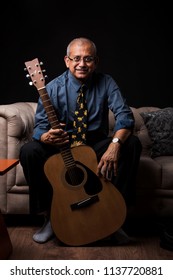 Senior Indian/asian Handsome Man Playing Or Posing With Musical String Instrument Like Guitar Or Violin, Standing Over Black Background Or Sitting On Sofa/couch