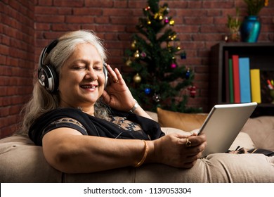 senior Indian/asian women listening to music using headphones while holding a tablet computer and sitting on sofa or couch at home
