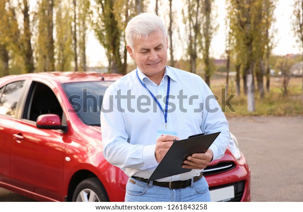 Senior instructor with clipboard near car\
outdoors. Get driving\
license