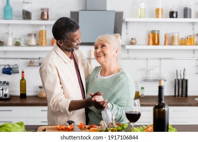 senior interracial husband and wife smiling while holding hands in kitchen - Powered by Shutterstock