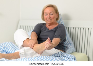 Senior lady massaging her bare foot to relive aches and pains as she sits relaxing against the pillows on her bed