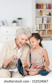 Senior Lady Teaching Her Granddaughter To Knit As They Relax Together On The The Sofa Laughing Together With Enjoyment