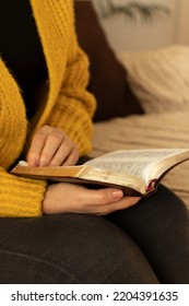 Senior Lady Wearing A Sweater Is Holding And Reading An Open Holy Bible Book With Golden Pages While Seating Peacefully On The Sofa At Home. Vertical Shot. A Close-up. Christian Study And Prayer.