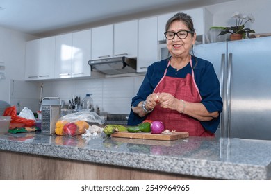 Senior Latina woman peeling garlic in the kitchen next to vegetables such as onion, pepper and tomato. The woman smiles while cooking - Powered by Shutterstock