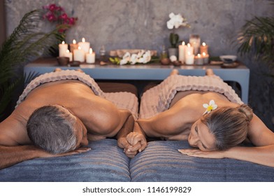 Senior loving couple lying naked at spa center holding hands. Mature woman and man lying on massage table for a spa treatment. Beautiful relaxed couple enjoy the peace after a beauty treatment.
