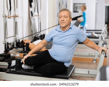 Senior male in activewear performing mermaid exercise on reformer Pilates machine for improving and maintaining mobility in rehabilitation center - Powered by Shutterstock