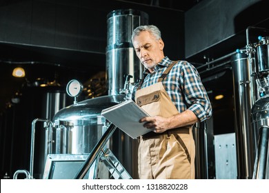 senior male brewer in working overalls looking at notepad while examining brewery equipment - Powered by Shutterstock