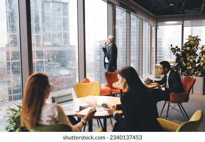 Senior male businessman speaking on phone while looking at skyscrapers and standing near window in modern office cafeteria near busy colleagues in formal clothes - Powered by Shutterstock