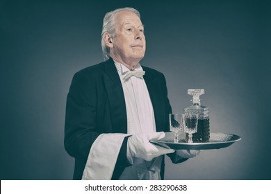 Senior Male Butler Wearing Tuxedo And Serving From Tray With Liquor Bottle And Crystal Cordial Glasses