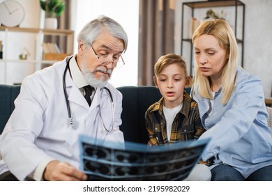 Senior Male Doctor, Sick Little Boy And His Mom Looking At X-ray Scan Image During Home Visit. Young Lady And Her Child Listening Their Doctor Explaining CT Scan And Talking About The Treatment.