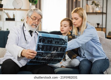 Senior Male Doctor, Sick Little Boy And His Mom Looking At X-ray Scan Image During Home Visit. Young Lady And Her Child Listening Their Doctor Explaining CT Scan And Talking About The Treatment.