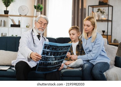 Senior Male Doctor, Sick Little Boy And His Mom Looking At X-ray Scan Image During Home Visit. Young Lady And Her Child Listening Their Doctor Explaining CT Scan And Talking About The Treatment.