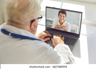 Senior Male Doctor Sitting At Desk In Hospital Office Using Laptop Computer Giving Video Call Consultation To Female Patient. General Practitioner Talking To Young Woman Online. View Over The Shoulder