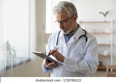 Senior Male Doctor Using Digital Tablet Computer Tech Providing Online Healthcare Telemedicine Ehealth Services Standing In Hospital Office. Serious Old Adult Physician Consulting Remote Patient.